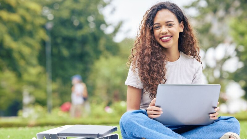 One young hispanic woman working on her laptop while sitting outside on an open field. A beautiful mixed race female student smiling while using her computer to study online on her university campus