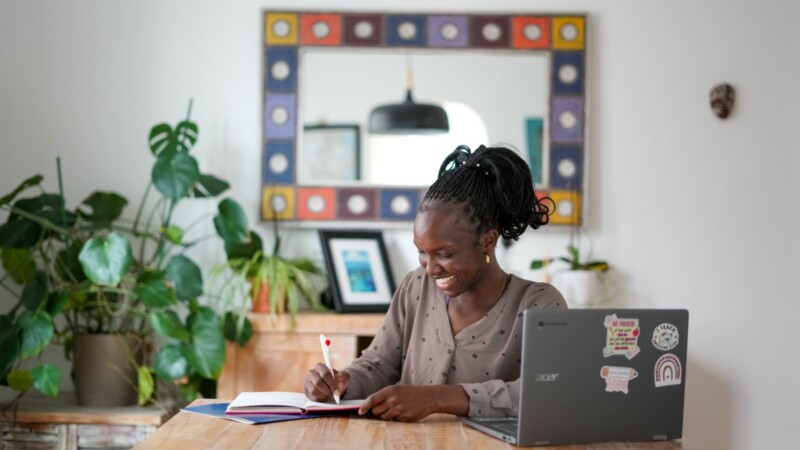 Smiling student sitting at a table writing in a notebook with a laptop on the table