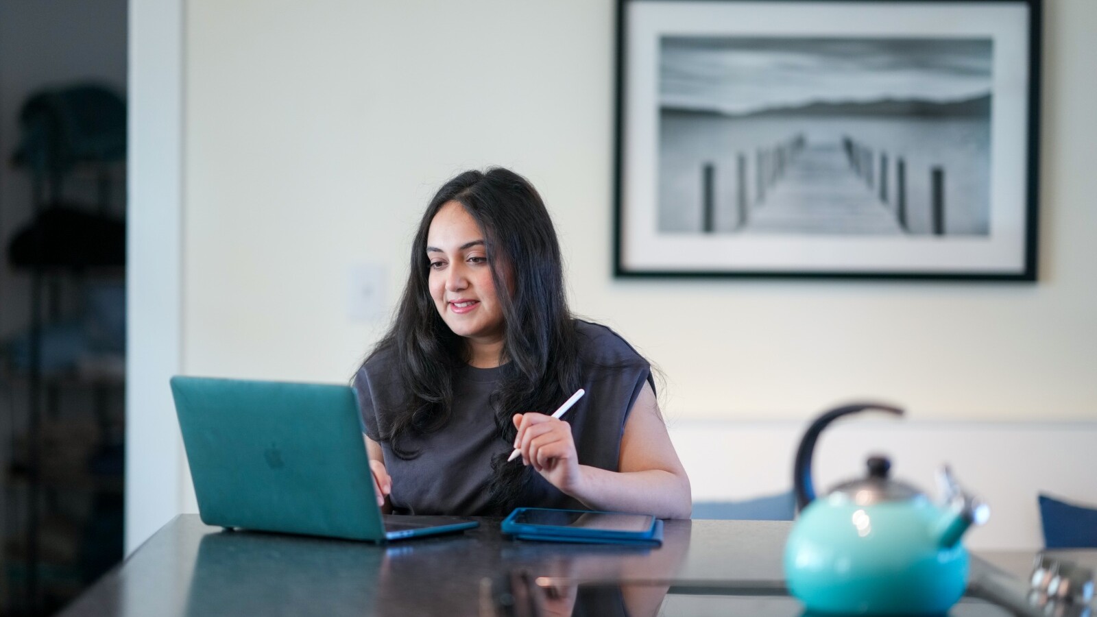 Student sitting at a kitchen counter working on a laptop with a kettle in the foreground