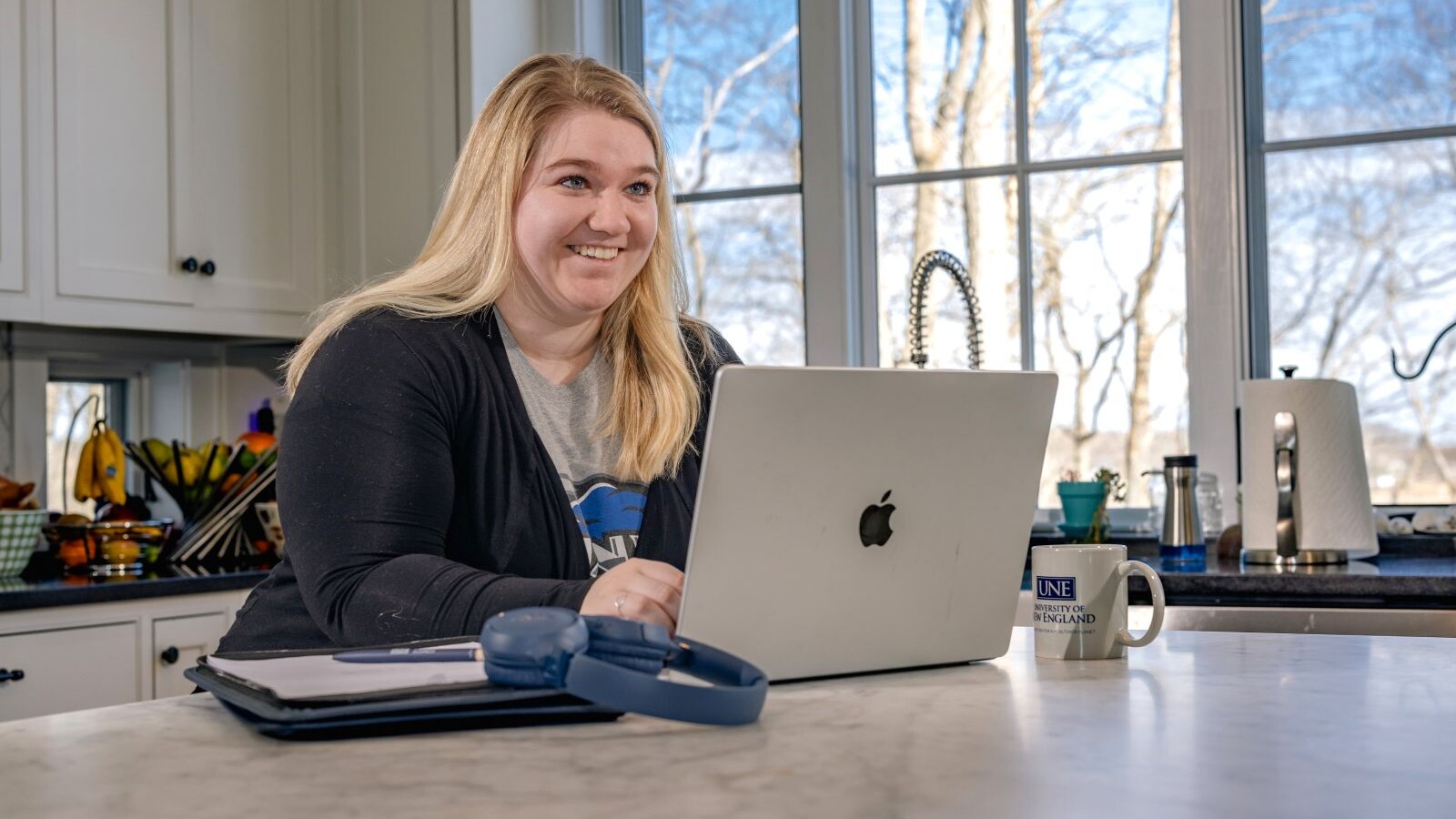 Smiling student sitting in a kitchen using a laptop with a mug and headphones nearby