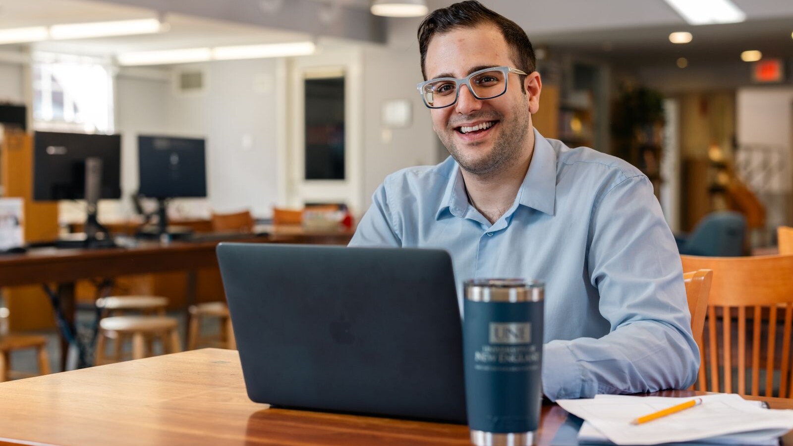 Student sitting at a desk in a large room typing on a laptop with a travel mug and notebook nearby