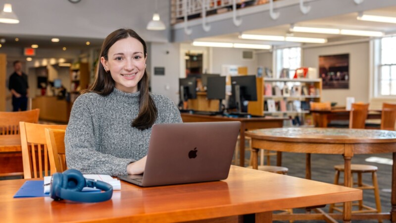 Student sitting at a table in a library using a laptop, with headphones and a notebook nearby