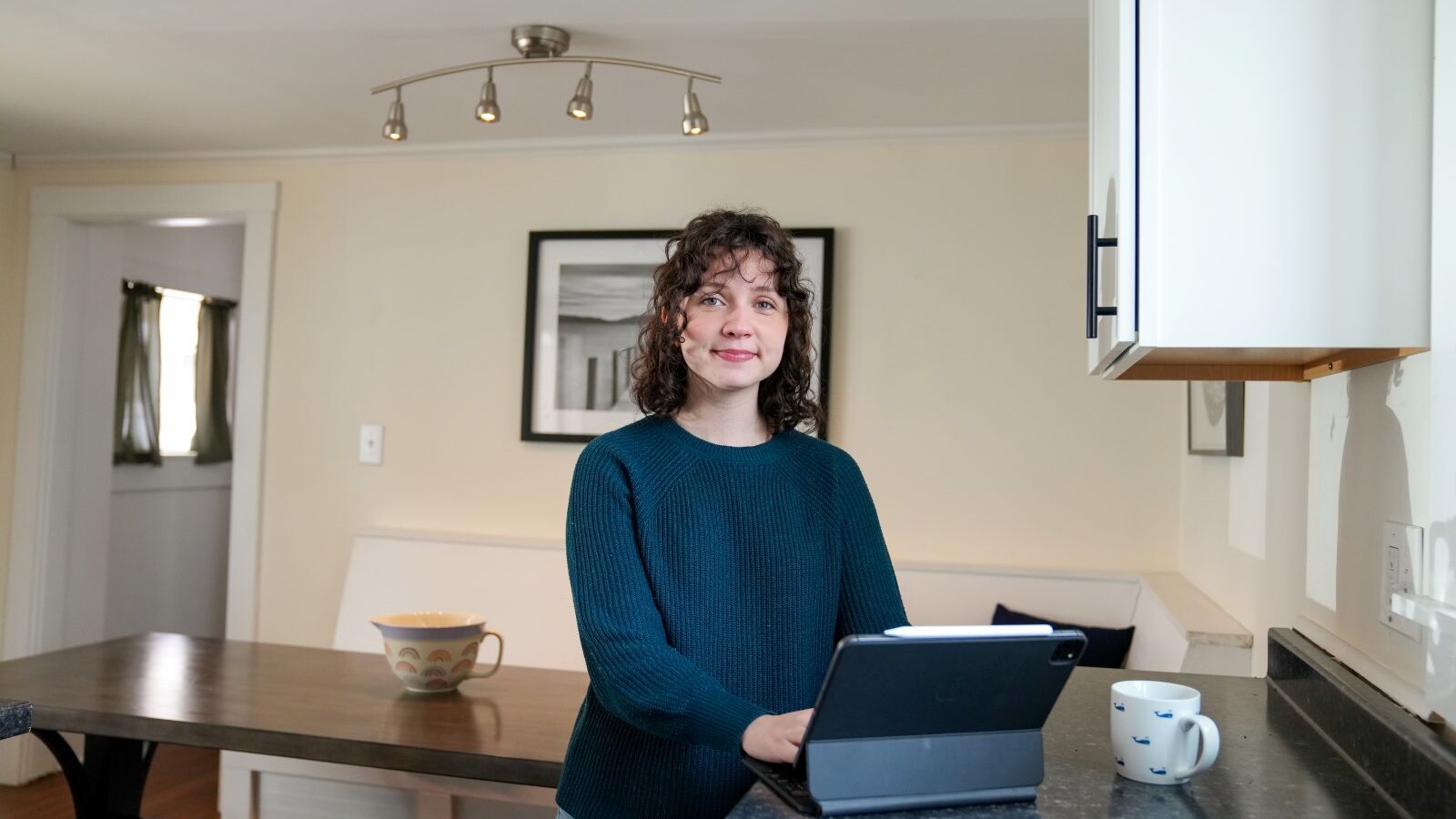 Student standing at a kitchen counter using a tablet with a mug nearby
