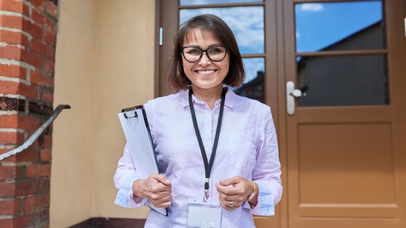 Portrait of female teacher, school worker looking at camera outdoor on steps of school building. Smiling positive woman counselor, school psychologist with clipboard. Education staff people concept