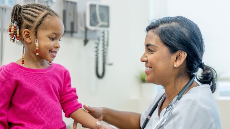 A caring South Asian nurse in scrubs greets a smiling young girl as she gently checks the child's arm during a pediatric clinic visit. Warm, trustworthy health care moment.