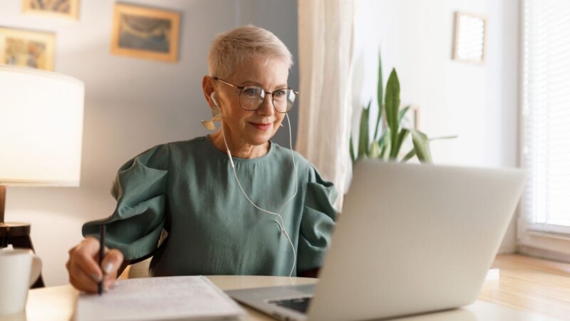 Indoor image of attractive stylish elegant woman in eyeglasses watching video tutorial in wired earphones, noting down main things in her textbook, sitting near window at table desk at home
