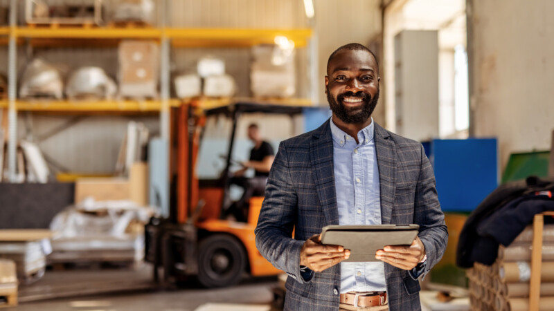 Confident entrepreneur standing in a warehouse with shelving units and forklift in background, holding a tablet. Showcasing leadership, logistics, and organizational skills in an industrial workplace.
