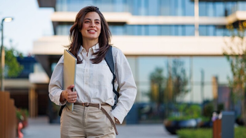 Happy college student walking outside modern campus building, carrying a laptop and backpack, looking forward