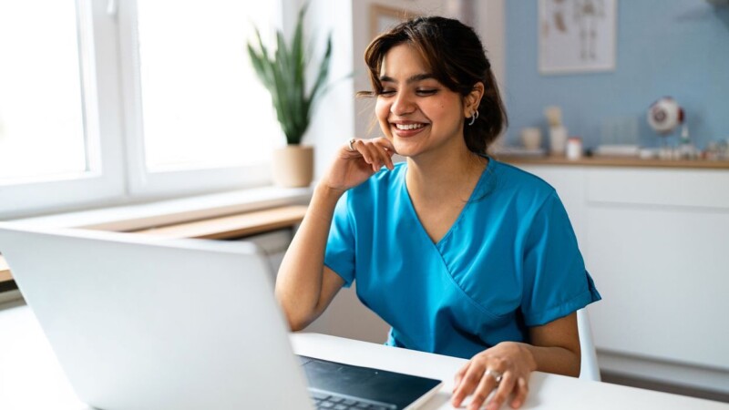 Female medical student of Indian ethnicity in blue medical scrubs, using a laptop at the modern ordination