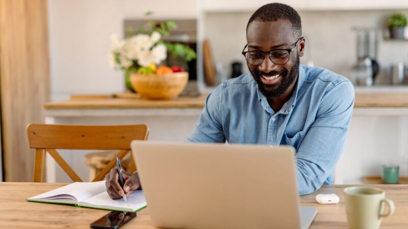 Happy man working from home taking notes looking at laptop
