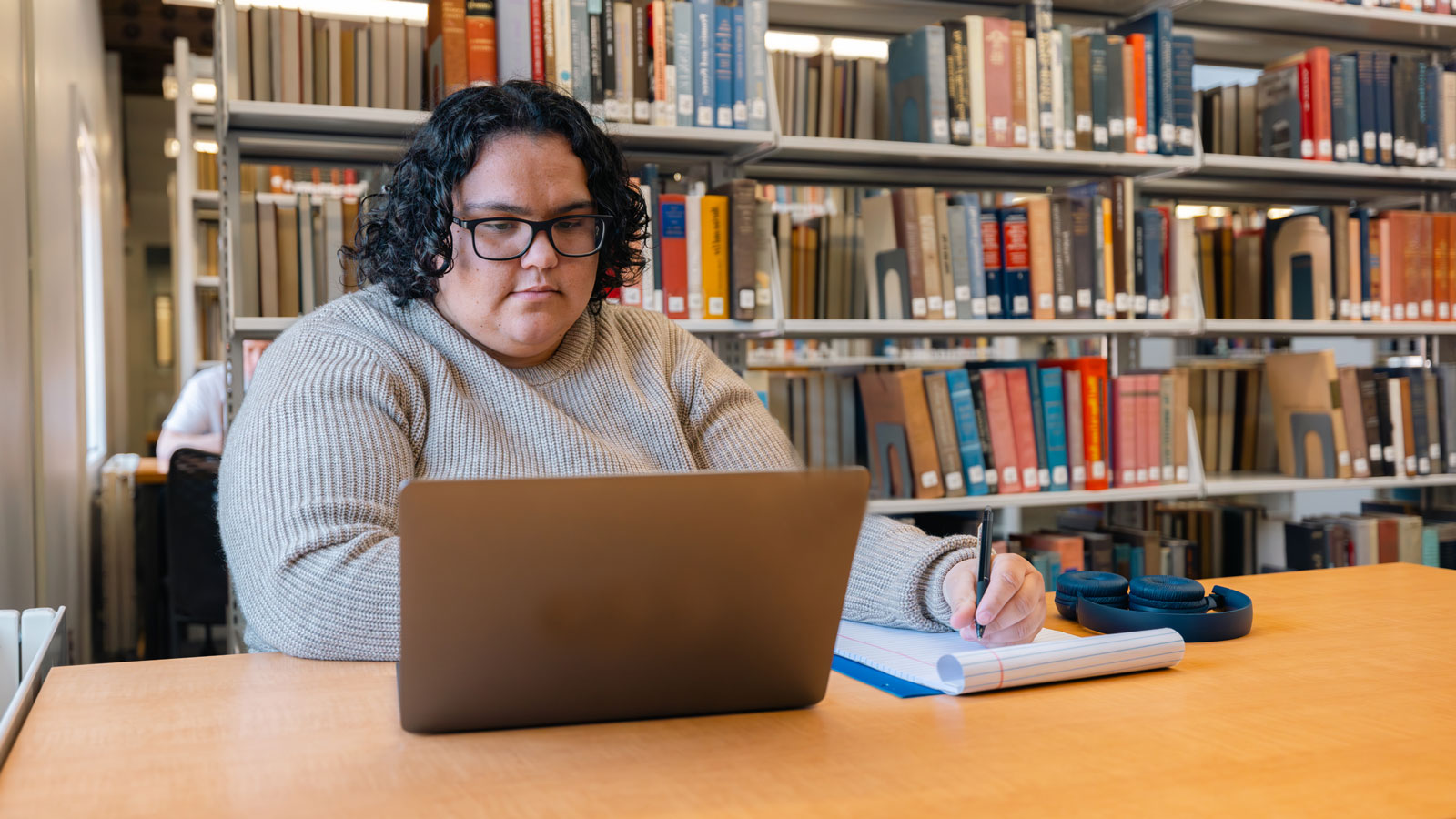 Student sitting at a desk in a library looking at a laptop and writing in a notebook