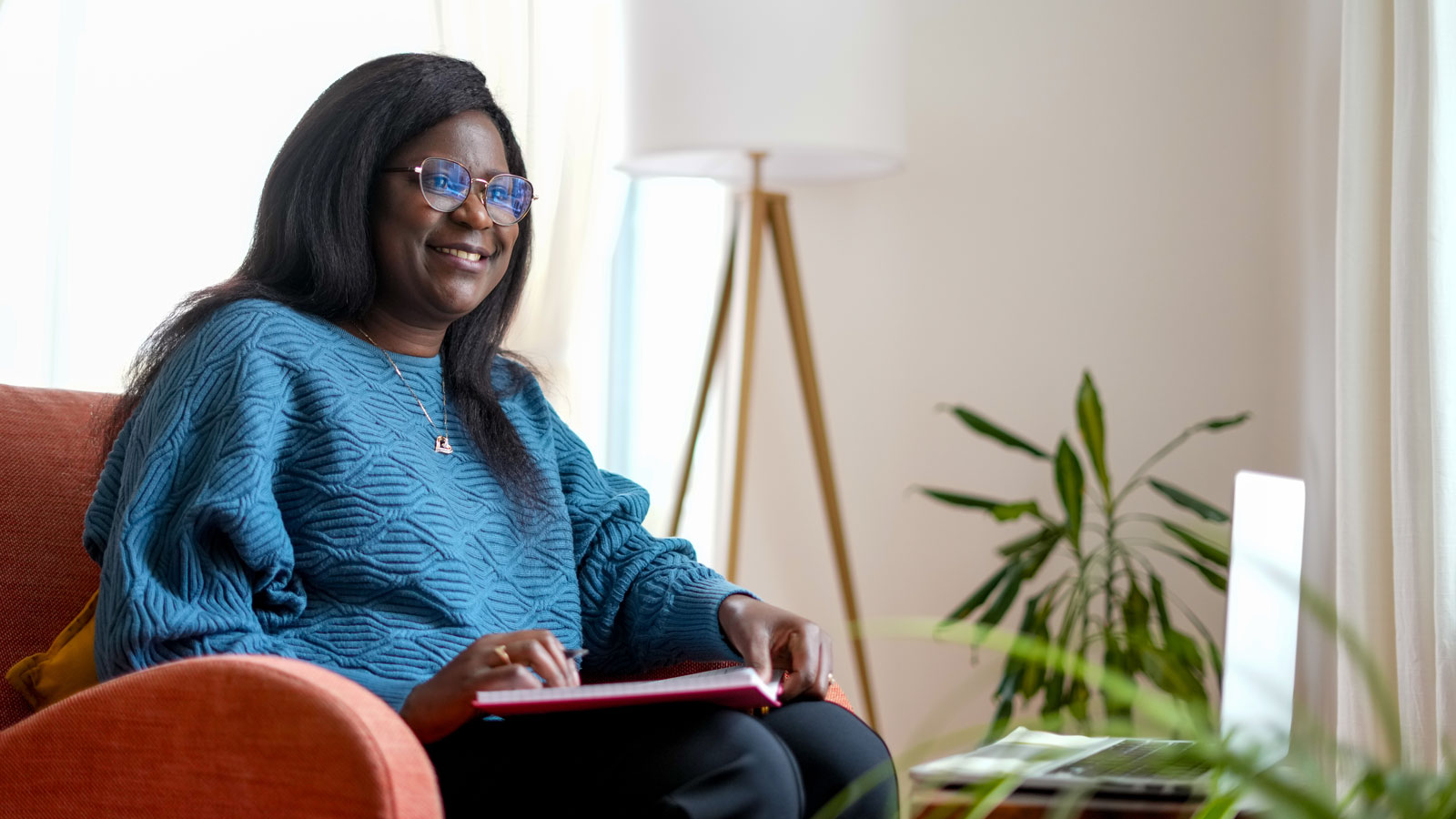 Student sitting in a chair holding a pen and an open notebook and smiling at a laptop