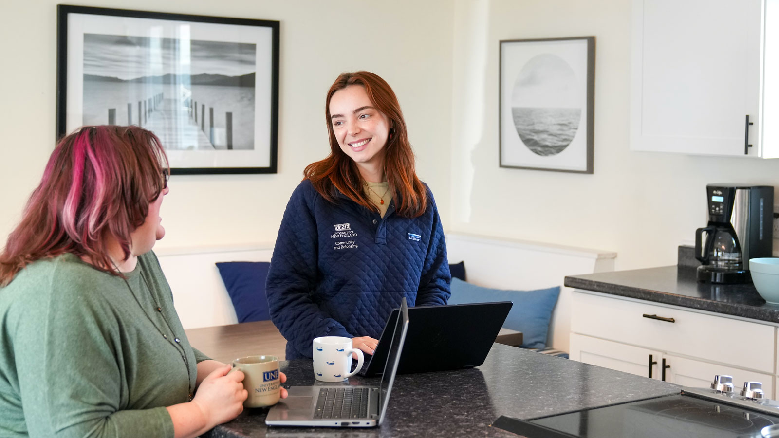 Two students talking at a kitchen counter with laptops and mugs