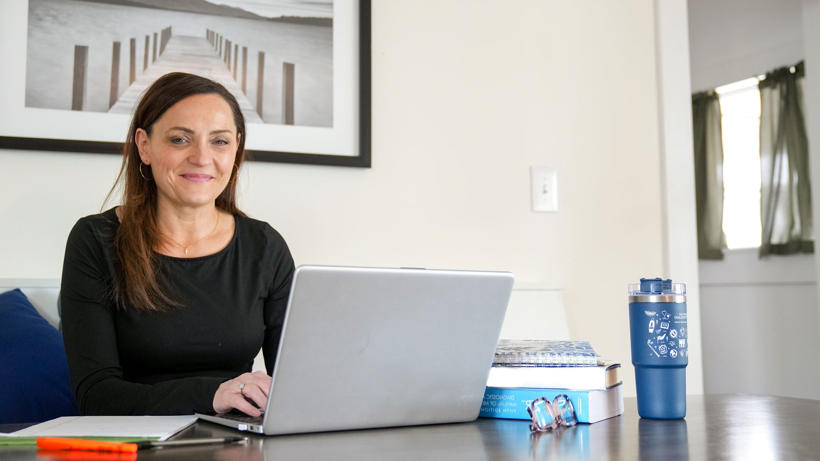 Smiling student sitting at a table, using a laptop, with a stack of books, glasses, notebook, and bottle nearby