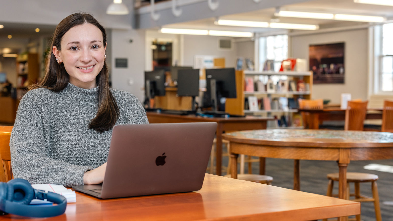 Student sitting at a table in a library using a laptop, with headphones and a notebook nearby